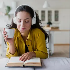 Mujer con auriculares y taza de café, disfrutando de audiolibros recomendados mientras lee un libro abierto.