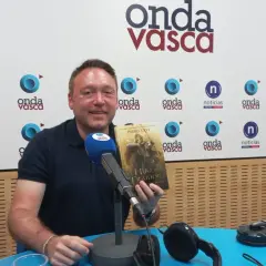 Hombre sonriente en estudio de radio, sosteniendo el libro "El Hijo del Traidor", como si recorriera el sendero del guardabosques orden.