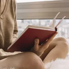Persona lee un libro de tapas rojas, sumergida en historias de novelas basadas en hechos reales mientras el sol entra por la ventana.