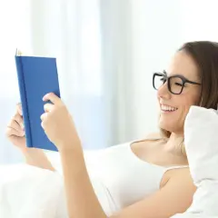 Mujer con gafas lee un libro azul en la cama, sonriendo. Una postura cómoda para leer.