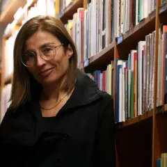 Una mujer con gafas sonríe entre estanterías llenas de libros, evocando la rica tradición de las escritoras catalanas famosas.
