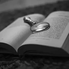 Un libro aperto con una lente d'ingrandimento posata sulle pagine, evocando un'atmosfera di mistero e segreti domestici, un vero domestic noir.