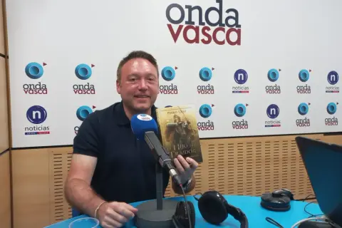 Hombre sonriente en estudio de radio, sosteniendo el libro "El Hijo del Traidor", como si recorriera el sendero del guardabosques orden.