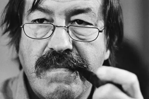 Primer plano de un hombre con gafas y bigote tupido, sosteniendo un bolígrafo. Podría ser uno de los escritores alemanes actuales.