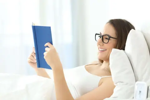 Mujer con gafas lee un libro azul en la cama, sonriendo. Una postura cómoda para leer.