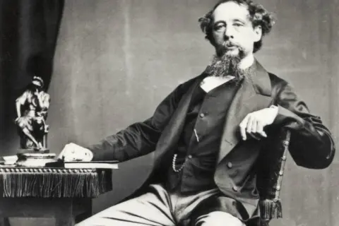 Charles Dickens, autor de libros famosos, posa con una estatua y un libro a su lado.
