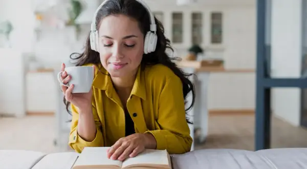 Mujer con auriculares y taza de café, disfrutando de audiolibros recomendados mientras lee un libro abierto.