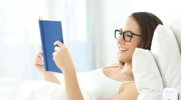 Mujer con gafas lee un libro azul en la cama, sonriendo. Una postura cómoda para leer.