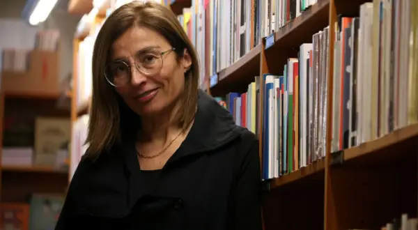 Una mujer con gafas sonríe entre estanterías llenas de libros, evocando la rica tradición de las escritoras catalanas famosas.