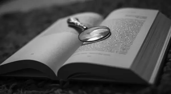 Un libro aperto con una lente d'ingrandimento posata sulle pagine, evocando un'atmosfera di mistero e segreti domestici, un vero domestic noir.