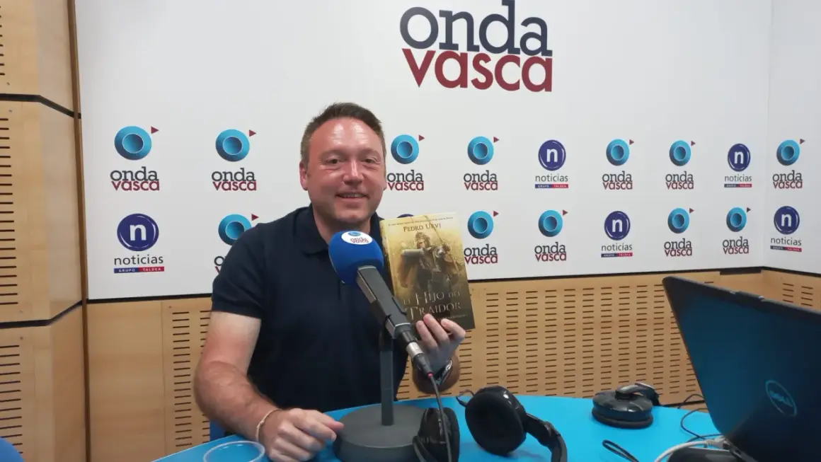Hombre sonriente en estudio de radio, sosteniendo el libro "El Hijo del Traidor", como si recorriera el sendero del guardabosques orden.