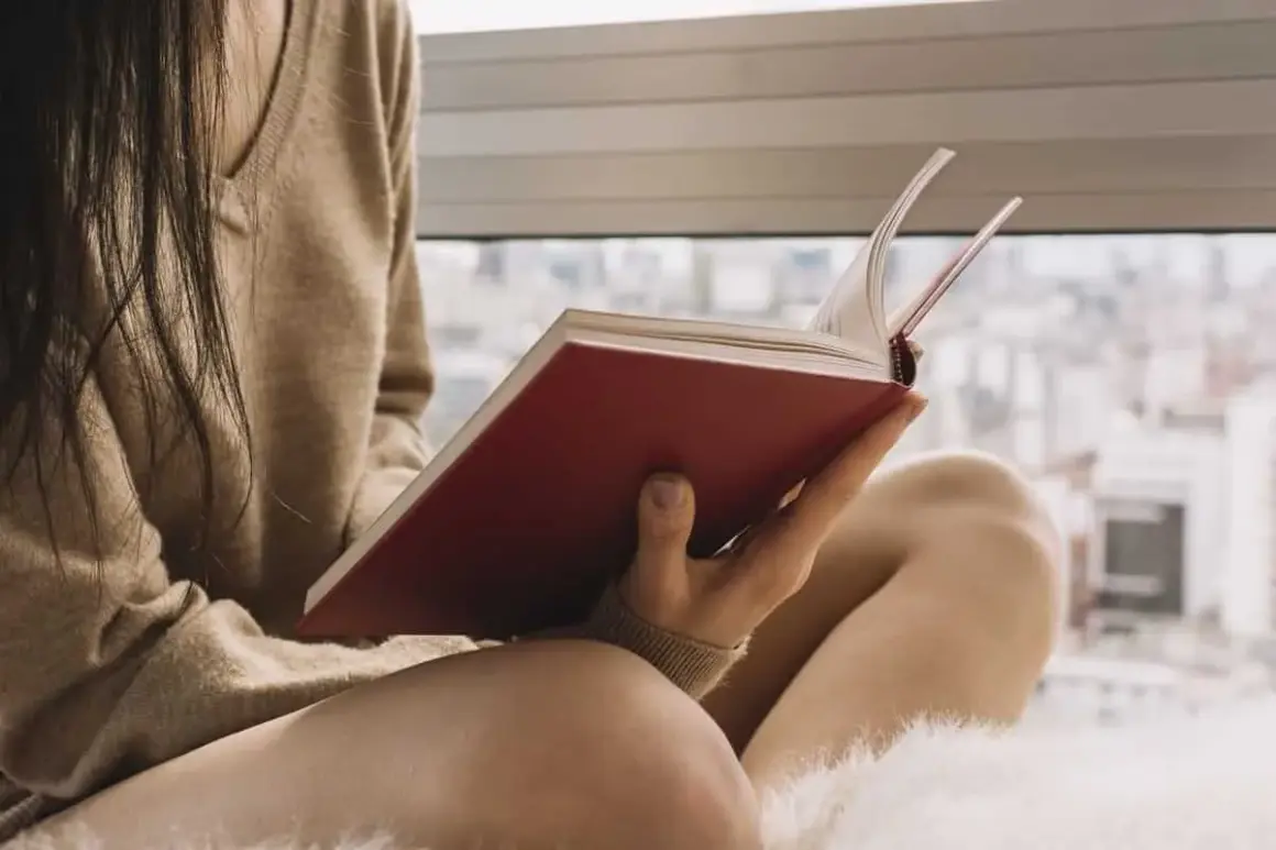 Persona lee un libro de tapas rojas, sumergida en historias de novelas basadas en hechos reales mientras el sol entra por la ventana.