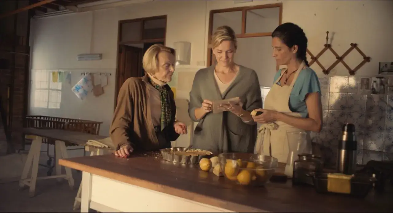 Tres mujeres en una cocina revisan una receta. En la encimera hay limones, leche y un molde con un pastel, quiz&aacute;s un pan de lim&oacute;n con semillas de amapola.