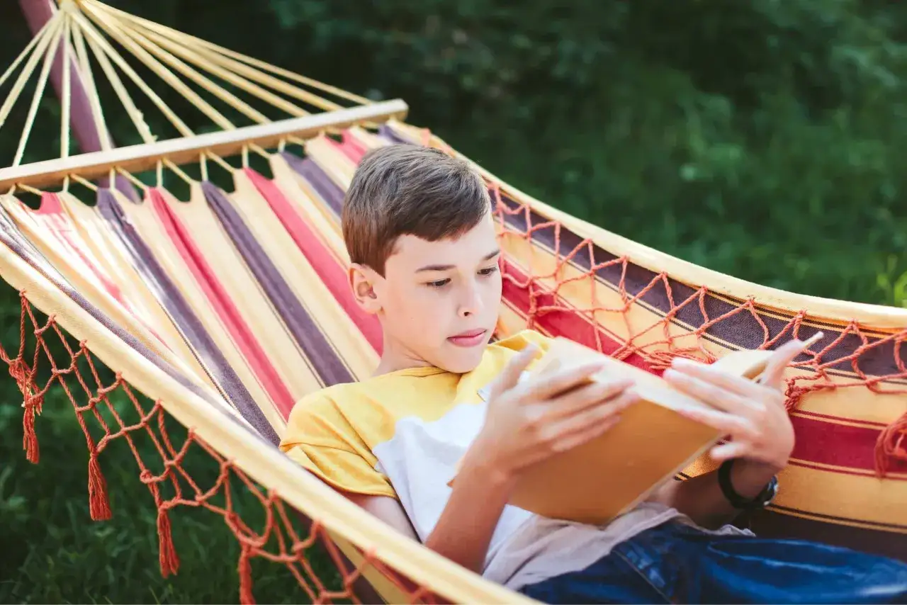 Un niño lee un libro en una hamaca a rayas, disfrutando de un momento de tranquilidad. ¡Perfecto para que leer este verano!
