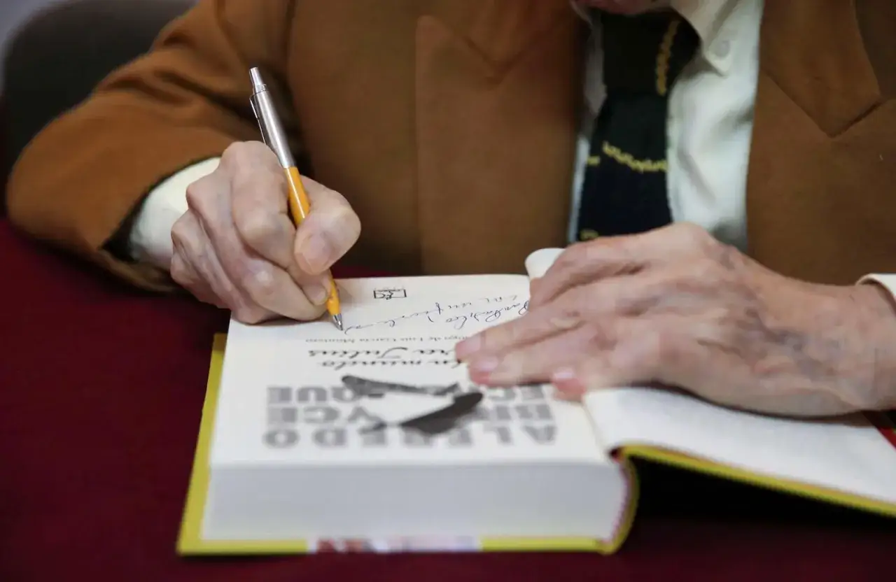 Un escritor italiano actual firma un libro. Sus manos, con un bolígrafo, escriben una dedicatoria en la página.