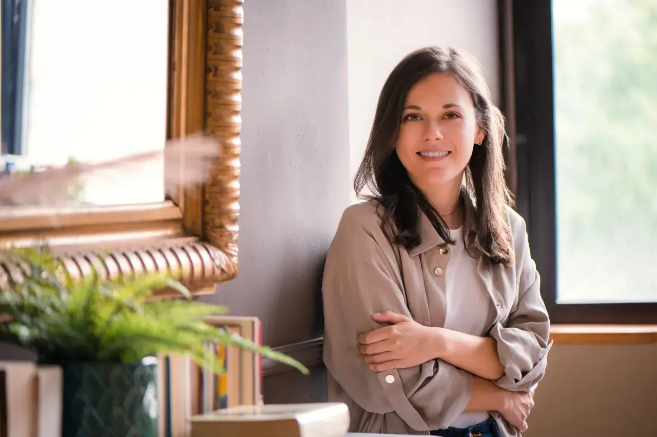 Mujer sonriente junto a un espejo y libros, lista para escribir su papel y tinta sinopsis.
