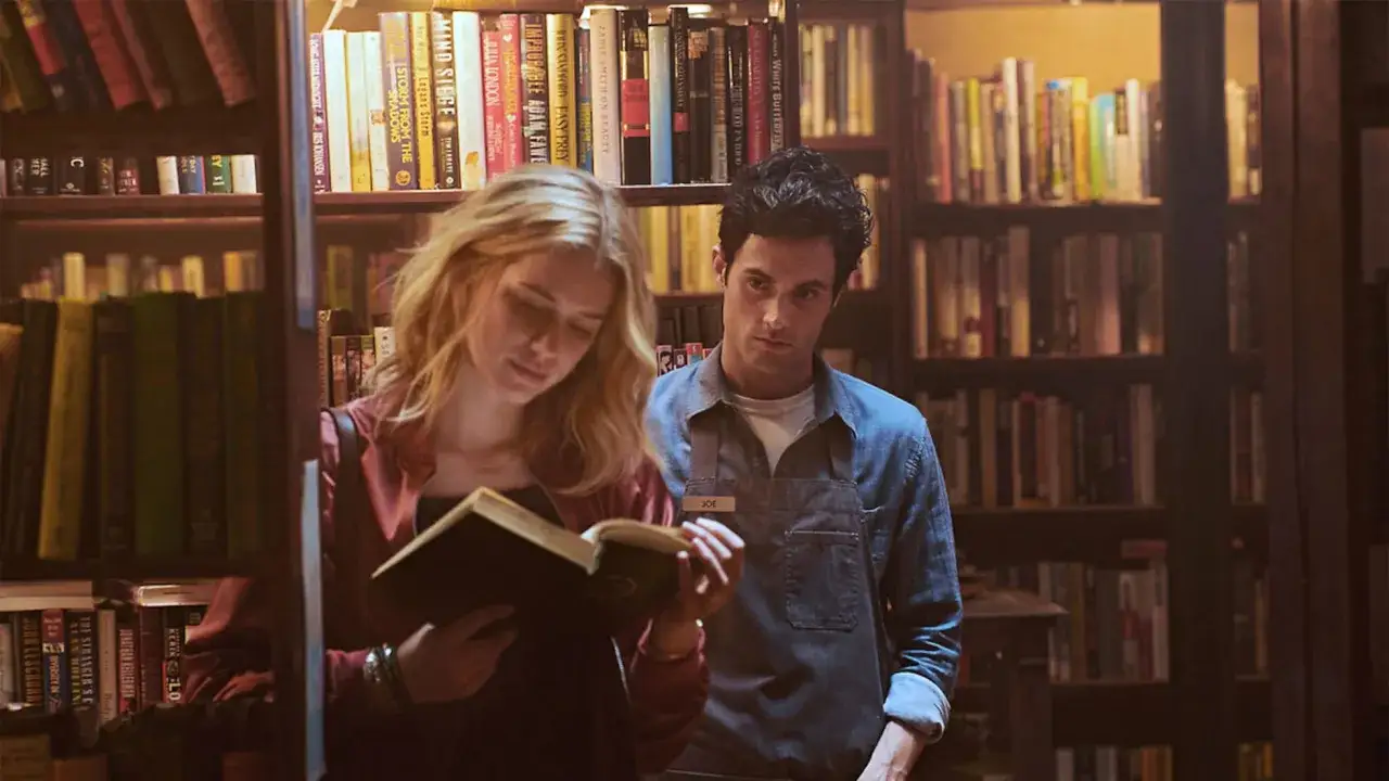Una mujer rubia lee un libro en una librer&iacute;a, mientras un hombre la observa. Escena de un oscuro libro de romance.