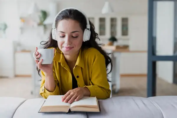 Mujer con auriculares y taza de café, disfrutando de audiolibros recomendados mientras lee un libro abierto.