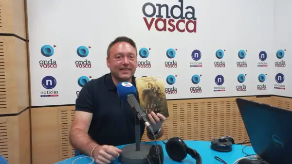 Hombre sonriente en estudio de radio, sosteniendo el libro "El Hijo del Traidor", como si recorriera el sendero del guardabosques orden.